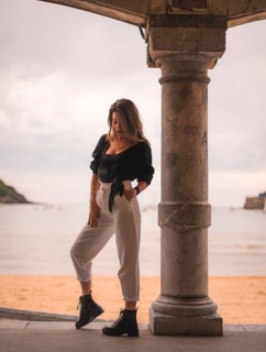 Lifestyle in the city with a blonde girl in white pants and a leather jacket near the beach. Photos next to a column in the shallows of the beach with his arm in his hair