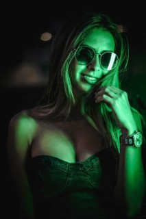 Caucasian brunette in a black dress illuminated by green led light reflected in black crystals. Urban night photography. Girl with sunglasses and saying quiet