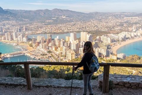 A young hiker girl with a hat looking at the city of Calpe from the path of the Penon de Ifach Natural Park