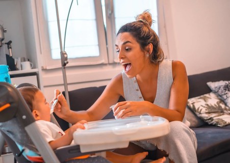 Young Caucasian mother feeding her son a yogurt while sitting in the highchair. Teleworking and caring for your child