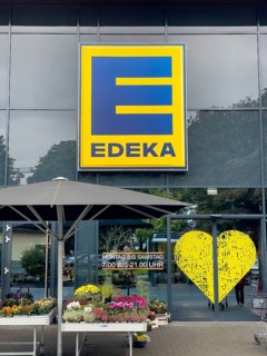 Logo with letter and lettering above entrance to supermarket below note on opening hours Monday to Saturday 7.00 a.m. to 9.00 p.m. to the right stylised heart of retail chain supermarket chain Edeka f...
