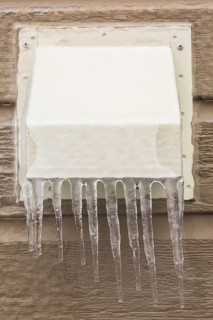 Metal exhaust vent covered with ice and icicles on tan clapboard wall of house
