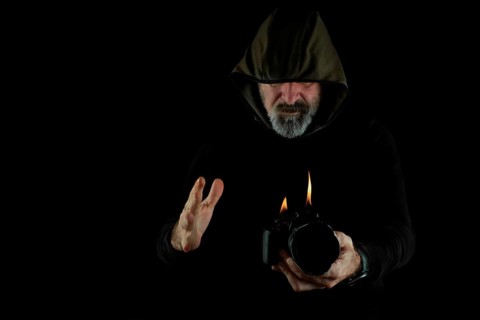 Man dressed in black with hood holding a burning reflex camera on a black background