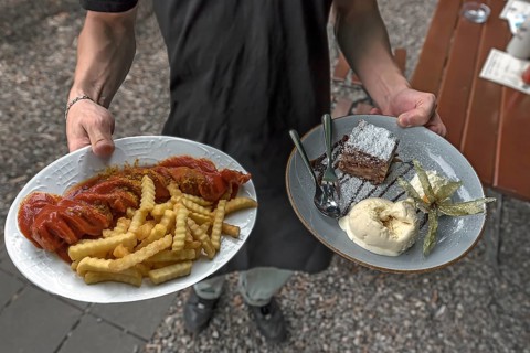 A waiter serves currywurst with fries and apple strudel with ice cream in a garden restaurant
