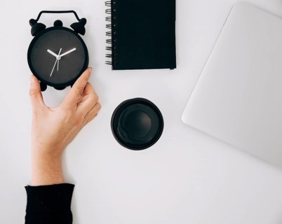 Close up person s hand holding black alarm clock spiral notepad laptop takeaway coffee cup white desk