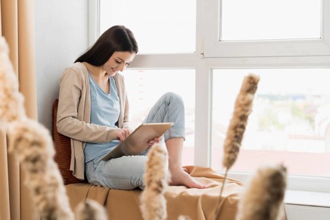 Side view woman sitting window working tablet