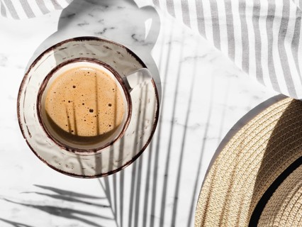 White cup with red shapes filled with creamy coffee white background with striped grey white cloth covered by ficus leaf shadow hat