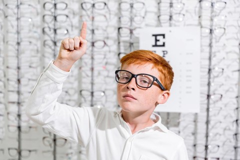 Boy with spectacle pointing upward direction standing against eyeglasses display background