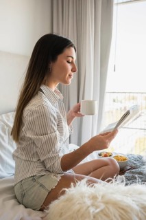Young woman sitting bed reading newspaper while drinking coffee