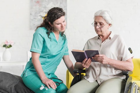 Disabled senior female patient sitting wheel chair reading book with nurse