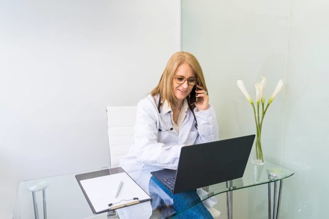 Smiling female doctor talking on the phone to a patient while looking at her medical records on laptop in medical office