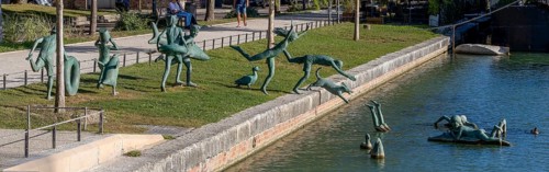 Panoramic photo of a group of bronze sculptures of playful children La Ribambelle Joyeuse by Belgian artist Tom Franzten on the banks and in the Canal du Trevois