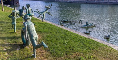 Panoramic photo of a group of bronze sculptures of playful children La Ribambelle Joyeuse by Belgian artist Tom Franzten on the banks and in the Canal du Trevois
