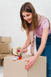 Smiling young woman holding packing machine sealing cardboard boxes with duct tape