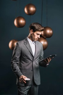 Young man holding disposable coffee cup looking digital tablet standing front streamlined mirror round copper chandelier