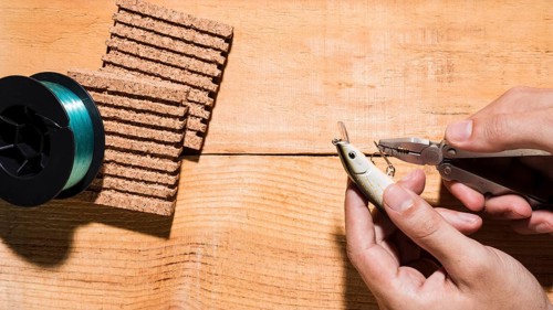 Close up man fixing hook with plier near reel cork board wooden desk