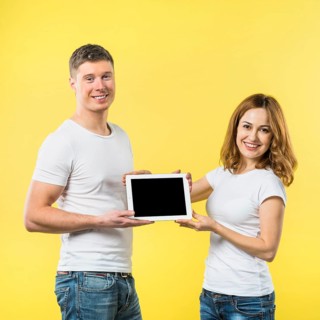 Portrait two happy young couple showing black screen digital tablet against yellow backdrop