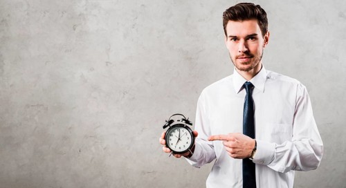 Portrait young businessman pointing black alarm clock standing against grey concrete wall