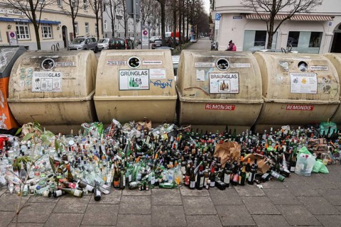 Overflowing recycling glass bottle containers with rows of bottles lined up in front on the footpath