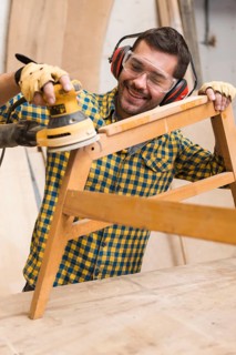 Smiling male carpenter softening edges wooden furniture with sander