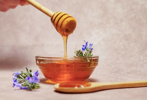 Woman holding a wooden spoon dripping honey over a glass bowl with fresh rosemary branches in bloom