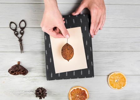 Elevated view hand making greeting card with dried leaf pinecone citrus slice lotus pod table