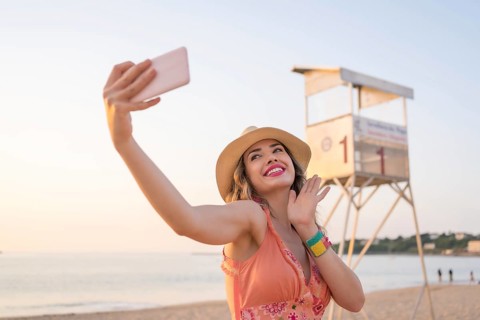 Woman with summer clothes waving at camera taking a selfie with the mobile on the beach during sunset