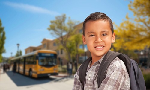 Cute young hispanic boy wearing a backpack near a school bus on campus