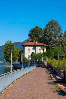 Walkway with Lamps and Railing and a House on the Waterfront on Lake Lugano with Mountain and Trees in a Sunny Summer Day in Porto Ceresio