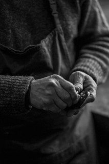 Monochrome photo of hands of Senior violinmaker wearing apron holding a tool chisel in workshop in Cremona