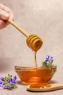 Woman holding a wooden spoon dripping honey over a glass bowl with fresh rosemary branches in bloom