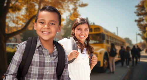 Happy young hispanic boy and girl wearing backpacks near a school bus on campus