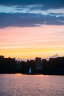 A sailing boat with a white sail lies on the water at sunset against the glowing orange and blue evening sky with dark clouds
