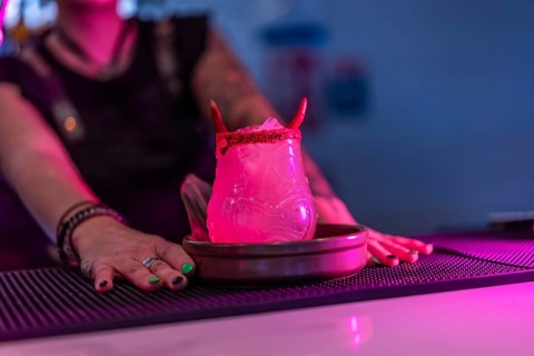 Bartender presenting a margarita cocktail decorated by devil horns in the counter of a night bar with neon lights