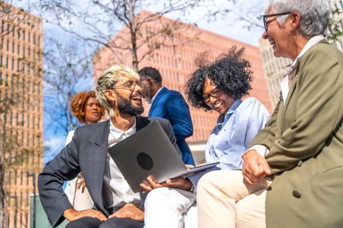 Smiling business team using laptop sitting outdoors in a bench outside a building