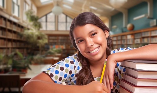 Happy young hispanic girl with a pencil and a stack of books doing homework in a library