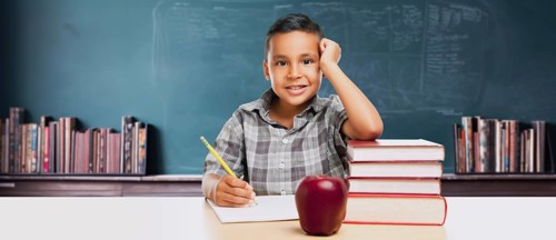 Happy young hispanic school boy at desk with books and apple in front of chalkboard