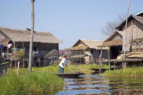 Intha man in proa at lakeside village with traditional bamboo houses on stilts in Inle Lake