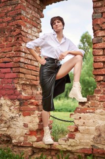 Image of a stylish beautiful woman in a white shirt and leather skirt in a park against the background of a destroyed building. The concept of style and fashion