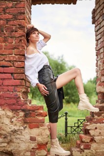 Image of a stylish beautiful woman in a white shirt and leather skirt in a park against the background of a destroyed building. The concept of style and fashion