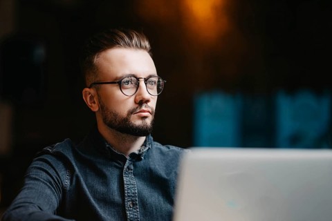 Young successful focused freelancer in a denim shirt working at a laptop in a covoking space and looking away