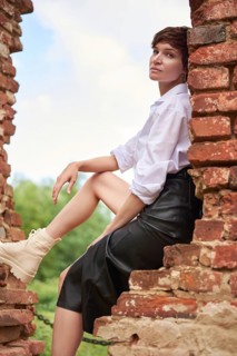 Image of a stylish beautiful woman in a white shirt and leather skirt in a park against the background of a destroyed building. The concept of style and fashion