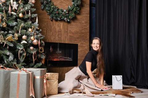 A joyful woman sits on the floor beside a warm fireplace decorated with a lush green wreath. The atmosphere is cozy and intimate