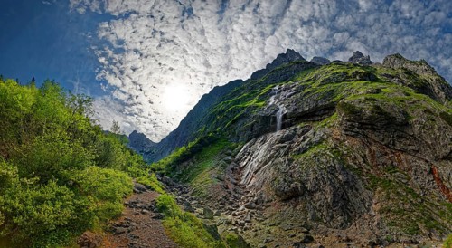 Sunlight breaks through the clouds over a rocky mountain landscape with lush greenery