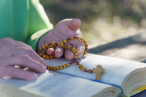 A woman's hands praying the rosary in the field over an open bible on a wooden table