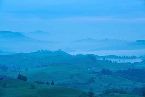 Moraine hill landscape with meadows and trees and some farms with morning fog and cloudy sky