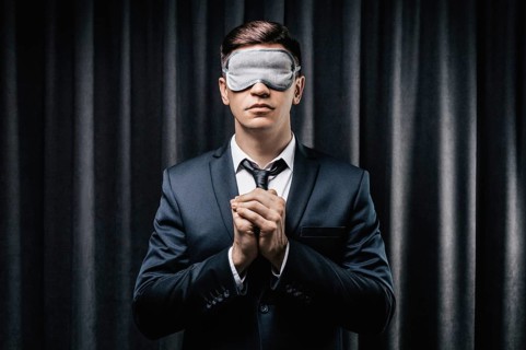Portrait of a man in a mask for sleeping. He prays against a black curtain. The concept of blind dates
