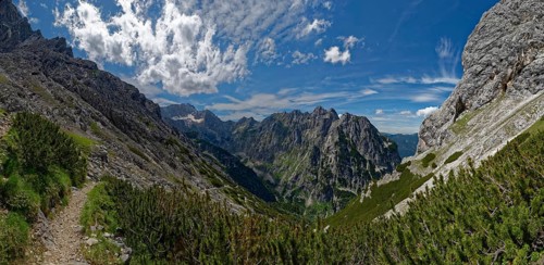 A narrow mountain path winds its way through a landscape of lush greenery and rock faces under a cloudy sky