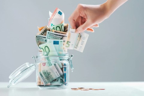 Female hand puts a bill into a glass piggy bank full of money. Thrift concept.