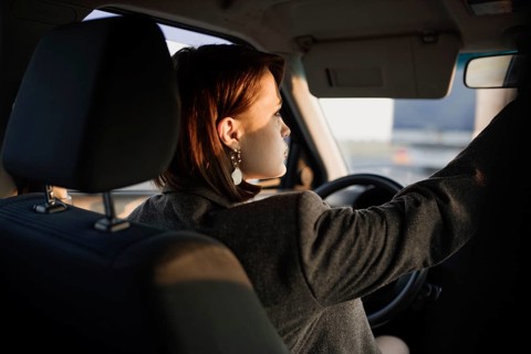 Young beautiful smiling woman taxi driver in a jacket sits behind the wheel of a car and adjusts the distant view mirror